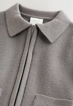 Pelēks ribots kardigans ar apkaklīti, priekšējo rāvējslēdzēja aizdari, divām kabatām un mīkstu teksturētu materiālu. Uz etiķetes ir uzraksts "NEXT baby."
