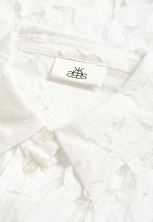 Weißes Spitzenhemd mit spitzem Kragen und zwei weißen Knöpfen, mit einem gestickten Muster und einem kleinen rechteckigen Markenetikett innen im Kragen.