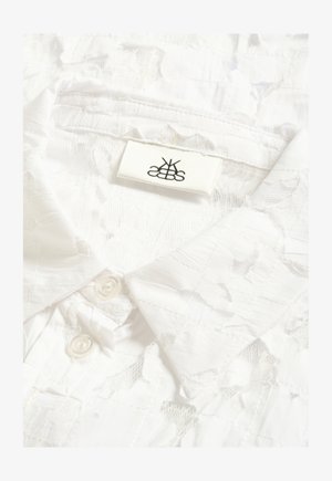 Valge pitsist särk terava krae ja kahe valge nupuga, millel on õmmeldud muster ja väikese ristkülikukujuline brändi sildike krae sees.