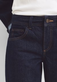 Jeans in denim blu scuro con cuciture arancioni, design a cinque tasche e chiusura con bottone in metallo. La superficie del tessuto appare liscia e robusta.