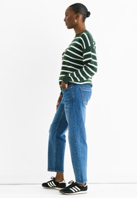 Grün-weißer gestreifter Strickpullover, blaue gerade Jeans und schwarze Sneakers mit weißen Streifen. Das Modell steht seitlich mit den Händen in den Taschen.