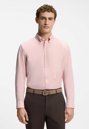 Jeune homme portant une chemise rose pâle boutonnée, rentrée dans un pantalon foncé avec une ceinture beige, se tenant devant un fond uni.