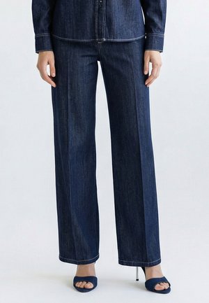 Frau trägt dunkelblaue, weit geschnittene Denim-Hosen kombiniert mit passenden, offenen High-Heel-Sandalen vor schlichtem Hintergrund.