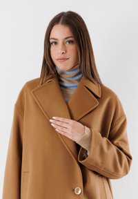 Cappotto di lana marrone con un design doppiopetto, ampi revers e un grande bottone. Indossato sopra un maglione a righe con collo alto.