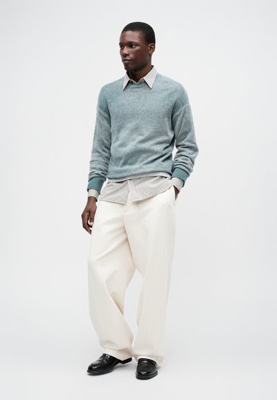 Helehelesin helesinine kootud sviiter valge ja tumesinise triibulise särke peal, koos kreemja laia säärega pükste ja mustade nahkkingadega.