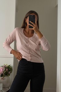 Gaiši rozā kardigāns ar apaļu kakla izgriezumu un pogu aizdari, izgatavots no mīkstas auduma. Saderīgs ar tumšajām džinsām. Līdzās ir ziedu kompozīcija.