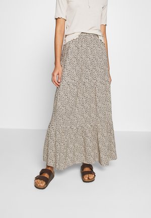 Mujer con una blusa de manga corta beige, falda larga color crema con motas negras y sandalias marrones de doble tira, de pie sobre un fondo liso.