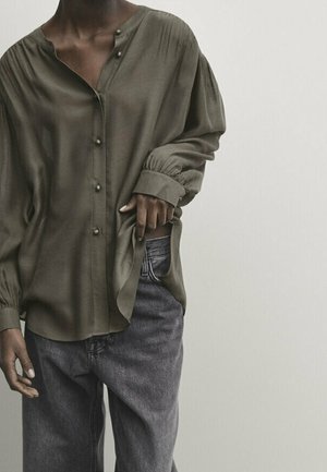 Persona dalla pelle scura che indossa una camicetta verde oliva con bottoni di perla e jeans denim grigio scuro larghi, con una mano che sistema l'orlo della camicia.