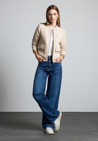 Veste beige matelassée avec deux poches avant, portée sur un haut blanc, associée à un jean évasé bleu foncé et des baskets blanches, sur un fond gris.