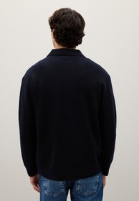 Maglione a maglia blu navy con colletto e vestibilità rilassata, dotato di maniche lunghe e una texture morbida, indossato sopra jeans azzurri.