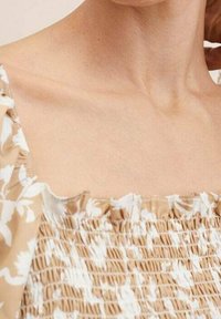 Haut à motifs floraux marron et blanc avec encolure à volants ; présente une texture élastique smockée, mettant en valeur une coupe ajustée.