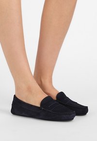 Mocassins en daim navy avec une texture lisse, présentant des coutures décoratives et une semelle plate, montrées sur des jambes du pied à la cheville.