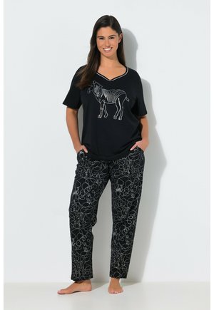 Smilende kvinde stående barfodet iført sort V-hals skjorte med hvid zebra-omrids og sorte bukser med hvide abstrakte og zebra designs.