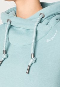 Ljusblå hoodie med en stor halsöppning, dragsko med metalländar och en slät, mjuk textur. Minimal logotyp är synlig.