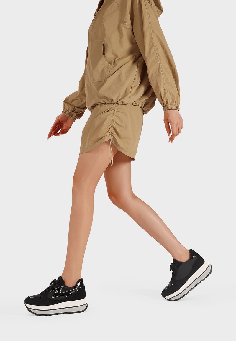 Persona che cammina in pantaloni corti arricciati color beige e giacca larga abbinata, indossando sneakers piattaforme bianche e nere su sfondo bianco.