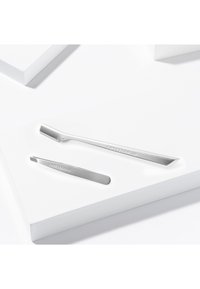 Pince à épiler fine en acier inoxydable inclinée et rasoir pour sourcils avec bords tranchants, posés sur une surface blanche.