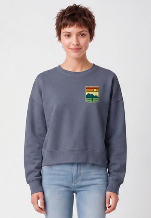 Jeune personne aux cheveux courts portant un sweat-shirt gris avec un motif coloré de montagne et de soleil avec l'inscription "Good Vibe" et un jean bleu clair.