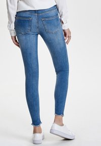 Jeans ajustados de denim azul, con una cintura de altura media, dobladillo deshilachado y dos bolsillos traseros. Combinados con zapatillas blancas.