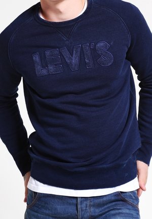 Felpa blu marino con logo Levi's testurizzato sul davanti, dotata di colletto e polsini a coste, indossata sopra una maglietta bianca.