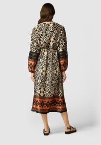Lång mönstrad klänning med intryckt midja, som har geometriska mönster i svart, beige och orange. Långa ärmar och glänsande tygtextur.