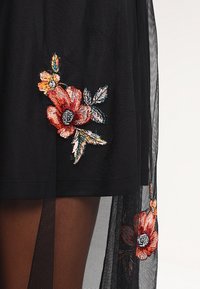 Tissu noir transparent avec des fleurs brodées rouges et orange et des feuilles vertes, superposé sur un matériau noir uni.