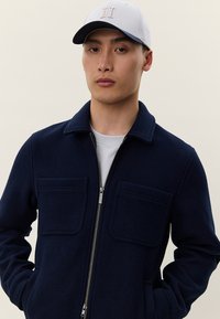 Ung mann iført en marineblå zip-up jakke med brystlommer og en hvit baseballcaps med et lite brodert design.