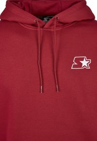 Sweat à capuche rouge avec une poche kangourou, des cordons de serrage, et un petit logo brodé blanc avec une étoile. Fabriqué à partir d'un tissu doux et durable.