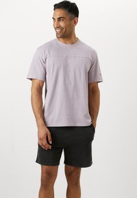 Light lavender short-sleeve t-shirt with a crew neckline and embroidered logo. Paired with dark shorts, showing a relaxed fit and texture.