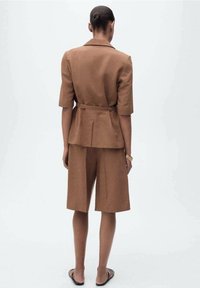 Giacca marrone su misura con maniche corte e pantaloni coordinati. Texture liscia, design strutturato, vita con cintura e dettaglio plissettato sul retro.