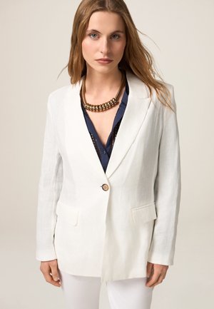 White linen blazer with notched lapels, two front pockets, and a single golden button. Worn over a navy blouse and paired with white pants.