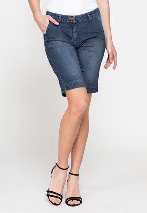Donna che indossa pantaloncini di jeans blu scuro fino al ginocchio e sandali neri con cinturini e tacco alto, in piedi con una mano in tasca.