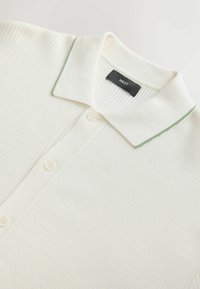 Polo blanc côtelé à trois boutons avec un col orné d'une fine bordure verte, étiquette indiquant la marque "NEXT".