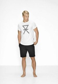 Hvid kortærmet t-shirt med et grafisk design i sorte striber, parret med sorte shorts; model står barfodet med hænderne i lommerne.