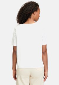 Hvid kortærmet T-shirt med firkantet halsudskæring og glat tekstur, bæres med beige bukser. Bagsiden er ensfarvet uden mønstre eller detaljer.