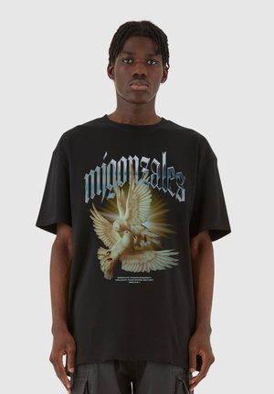 Sort bomulds t-shirt med et grafisk motiv af to duer i flugt. T-shirten har korte ærmer og en løs pasform, med fed tekst over designet.