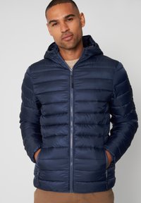 Giubbotto imbottito blu navy con cappuccio, caratterizzato da un design trapuntato, zip anteriore e due tasche laterali. Tessuto liscio e materiale leggero.