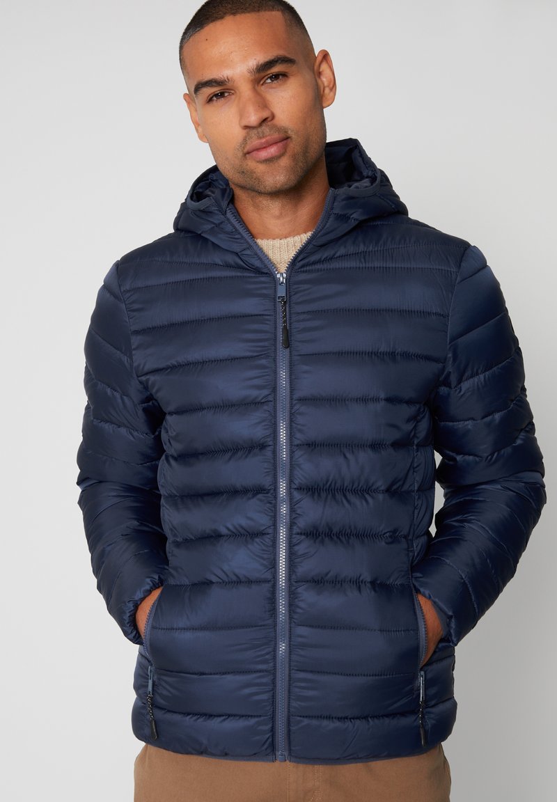 Giubbotto imbottito blu navy con cappuccio, caratterizzato da un design trapuntato, zip anteriore e due tasche laterali. Tessuto liscio e materiale leggero.
