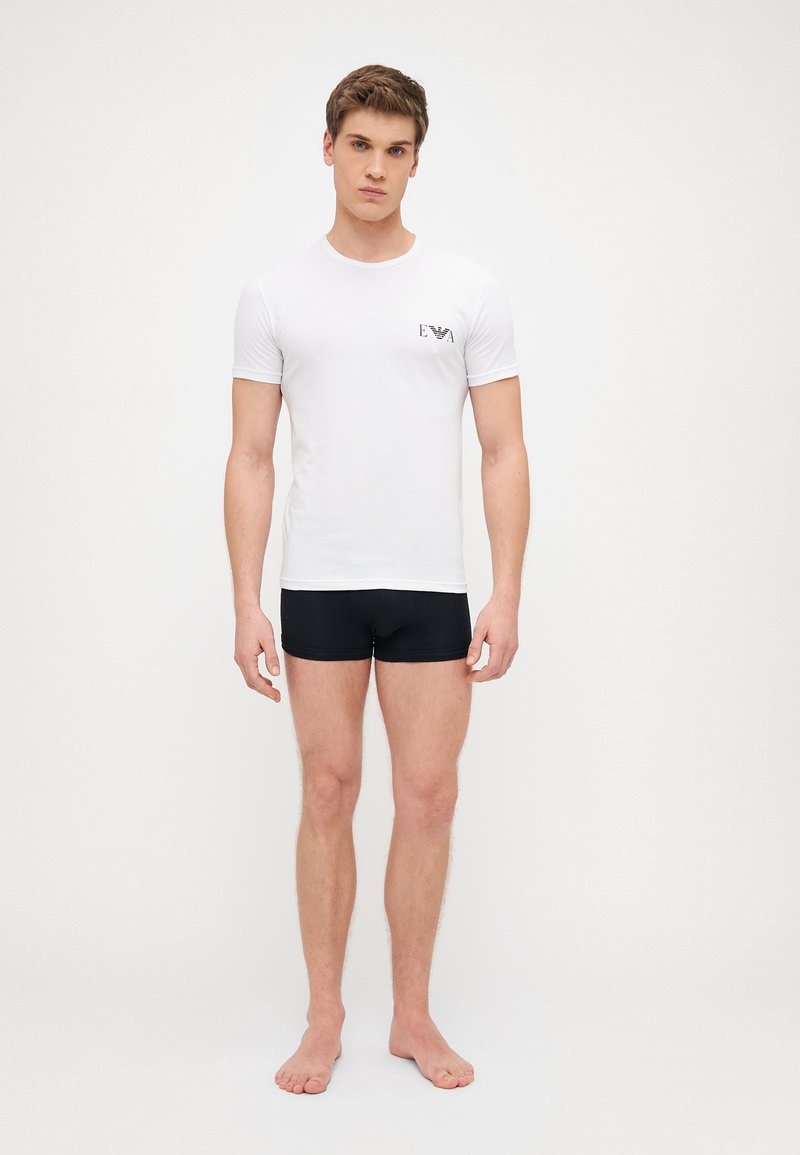 White short-sleeve T-shirt with a black logo on the left chest, paired with black fitted shorts. Smooth, lightweight fabric.