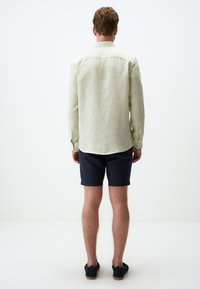 Hellgrünes Langarmhemd mit strukturiertem Muster, Knopfmanschetten und abgerundetem Saum, kombiniert mit marineblauen Shorts und dunklen Espadrilles.