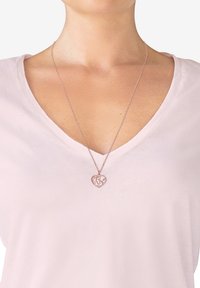 Hjerteformet vedhæng i rosaguld med globusdesign på en delikat kæde, båret over en lys pink V-hals top.