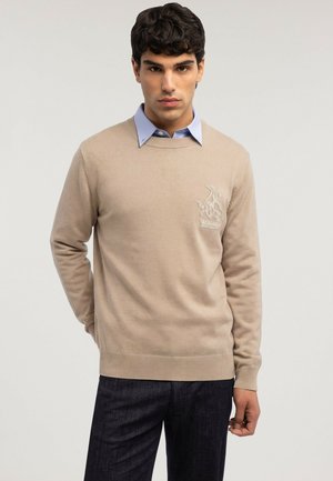 Mann trägt einen beigen Pullover mit Rundhalsausschnitt und gesticktem Logo über einem blauen Hemd mit Kragen sowie dunkler Hose vor neutralem Hintergrund.