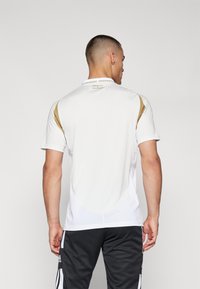 Camiseta deportiva blanca con detalles dorados en los hombros, mangas cortas y un dobladillo redondeado. Presenta un cuello y una tela texturizada.