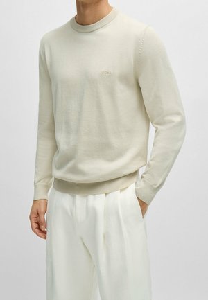 Homme portant un pull à manches longues de couleur crème avec un petit logo "BOSS" sur la poitrine, associé à un pantalon plissé blanc.