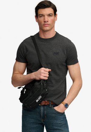 Mand iført en mørkegrå t-shirt og blå jeans holder en sort crossbody taske med "Superdry" logo, iført et brunt ur og bælte.