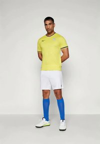 Gul fotbollströja med svarta detaljer, vita shorts och blå knälånga strumpor. Tröjan har ett subtilt mönster och en logo på bröstet.