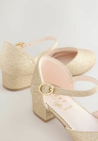 Børns gul glimmer hæle med ankelremme og oval spænder besat med rhinsten på en ensfarvet baggrund.