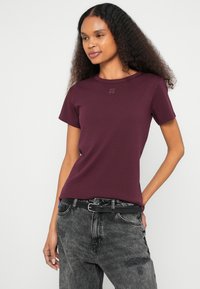 Μπορντό κοντομάνικο μπλουζάκι με στρογγυλή λαιμόκοψη και διακριτικό λογότυπο "HUGO" στο στήθος, συνδυασμένο με φθαρμένο γκρι τζιν και μαύρη ζώνη.