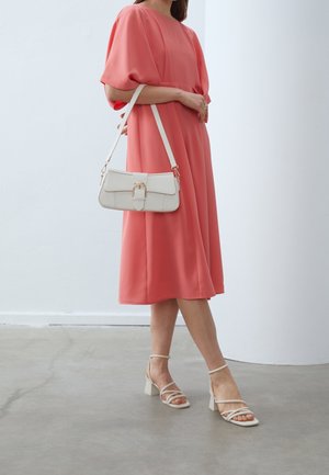 Femme en robe midi corail portant des sandales à talons blocs blanches à brides et un sac à main blanc avec boucle dorée sur l'épaule.