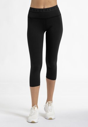 Leggings neri in tessuto elasticizzato con una ampia fascia in vita elastica che presenta il nome del marchio. Indossati con scarpe da ginnastica bianche.