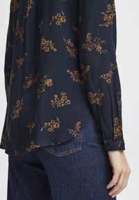 Marineblå bluse med blomstermønster i guld, med en let glans, lange ærmer og en buet kant, kombineret med mørke denimjeans.